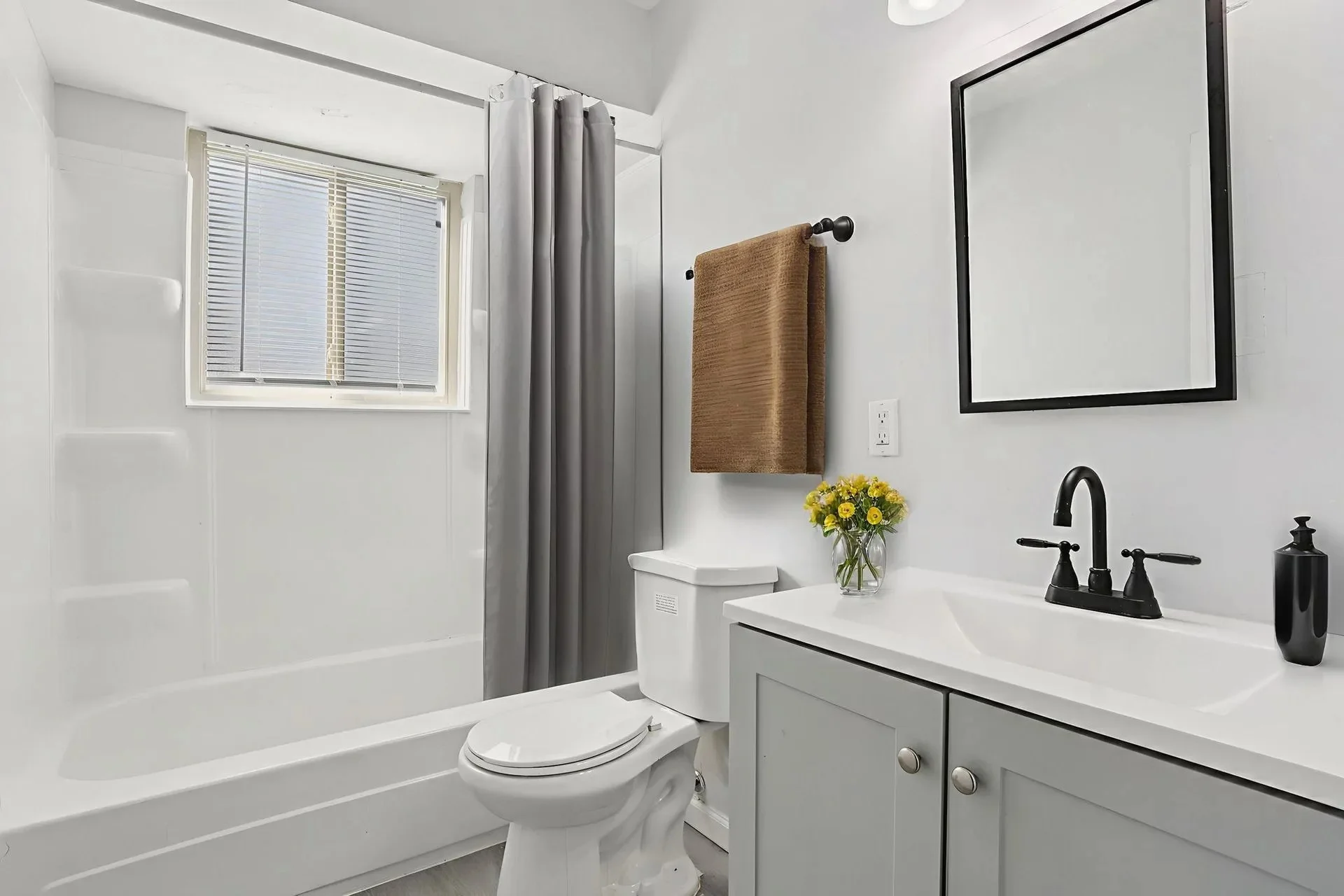 A clean and modern bathroom featuring a bathtub with shower, a white toilet, and a sink with two cabinets underneath. There is a mirror above the sink, a decorative towel hanging, and a vase of yellow flowers on the counter.