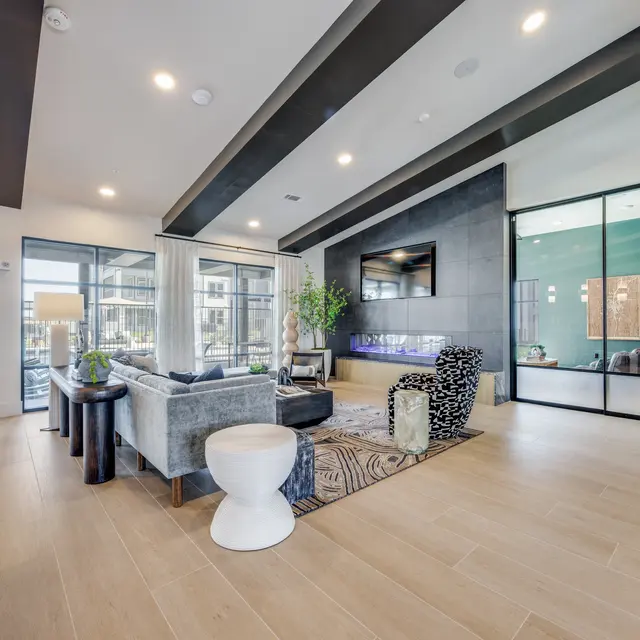 A modern living room featuring a grey sofa, white decorative table, and a large TV on the wall. The space is bright with large windows and elegant decor.