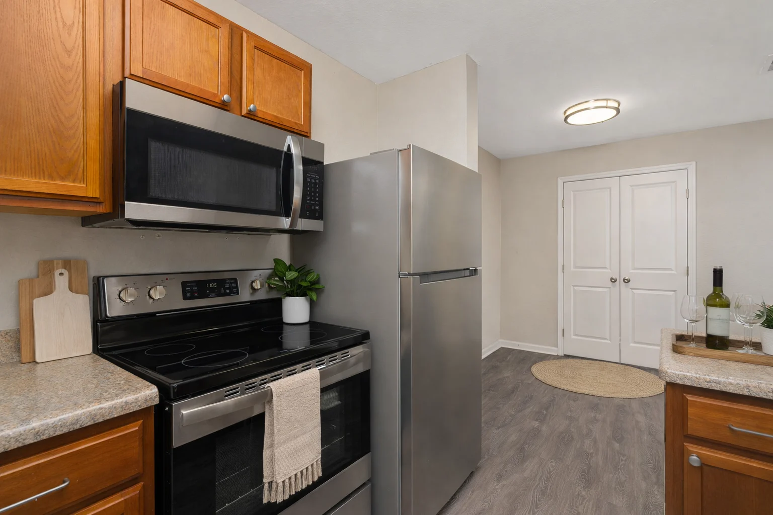 A modern kitchen featuring wooden cabinets, stainless steel appliances, and a gray floor. The kitchen includes an oven, microwave, and refrigerator. There is a cutting board and a plant near the stove, and a round rug near an entrance.