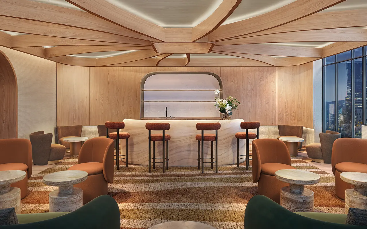 A stylish lounge area with a bar and contemporary seating. The bar, made of light-colored stone, is surrounded by high stools. The interior features wooden elements and various comfortable chairs in warm colors like orange and green. Soft lighting enhances the inviting atmosphere, complemented by a view of the city outside.