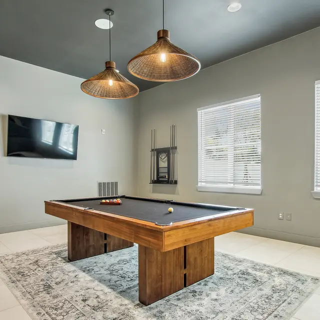 Chic Game Room with Pool Table A stylish game room featuring a wooden pool table on a decorative rug. The room is well-lit with modern pendant lights hanging from the ceiling and has a large flat-screen TV mounted on the wall. Windows with blinds allow natural light to enter, enhancing the contemporary feel.