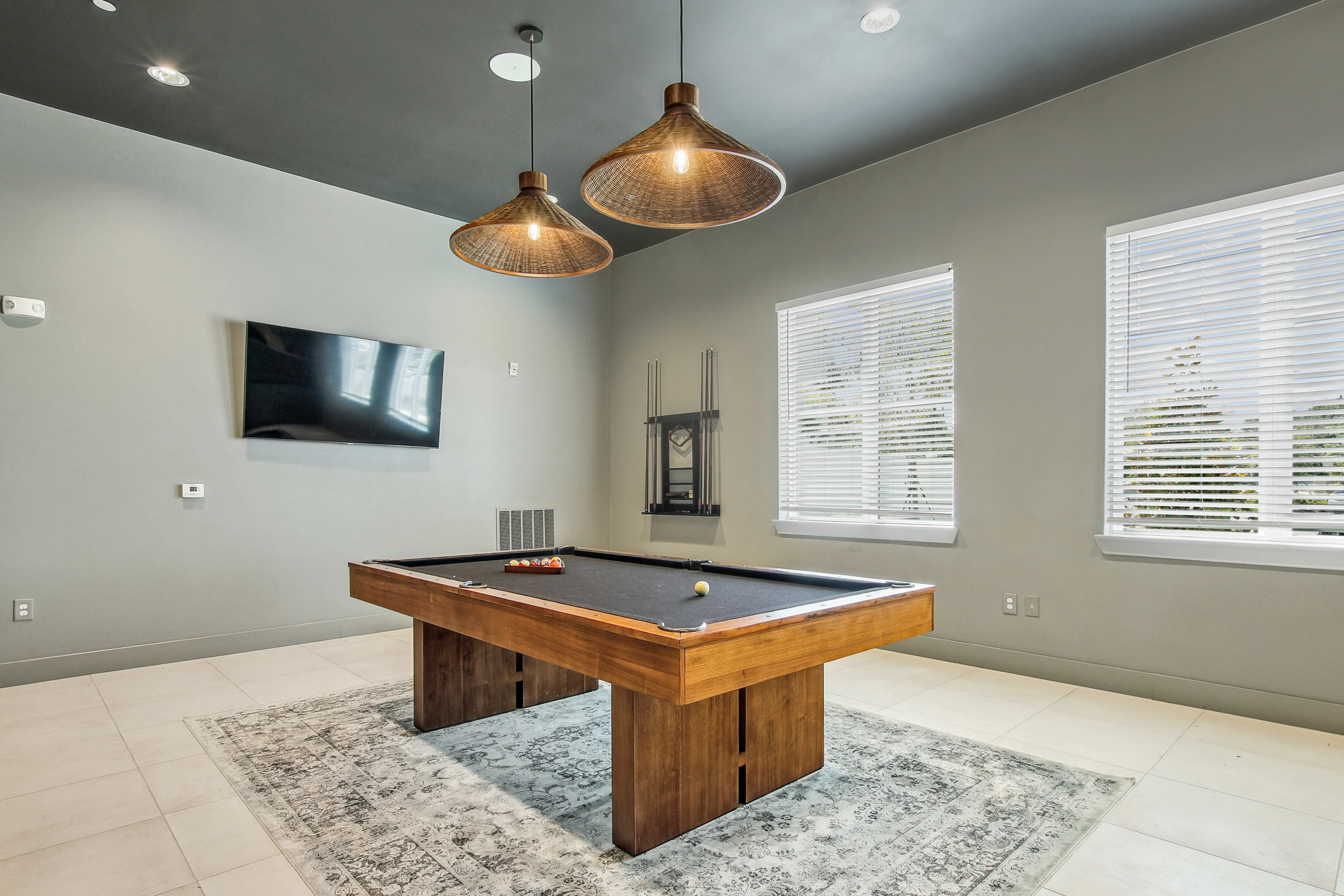 Sundance Trails A stylish game room featuring a wooden pool table on a decorative rug. The room is well-lit with modern pendant lights hanging from the ceiling and has a large flat-screen TV mounted on the wall. Windows with blinds allow natural light to enter, enhancing the contemporary feel.