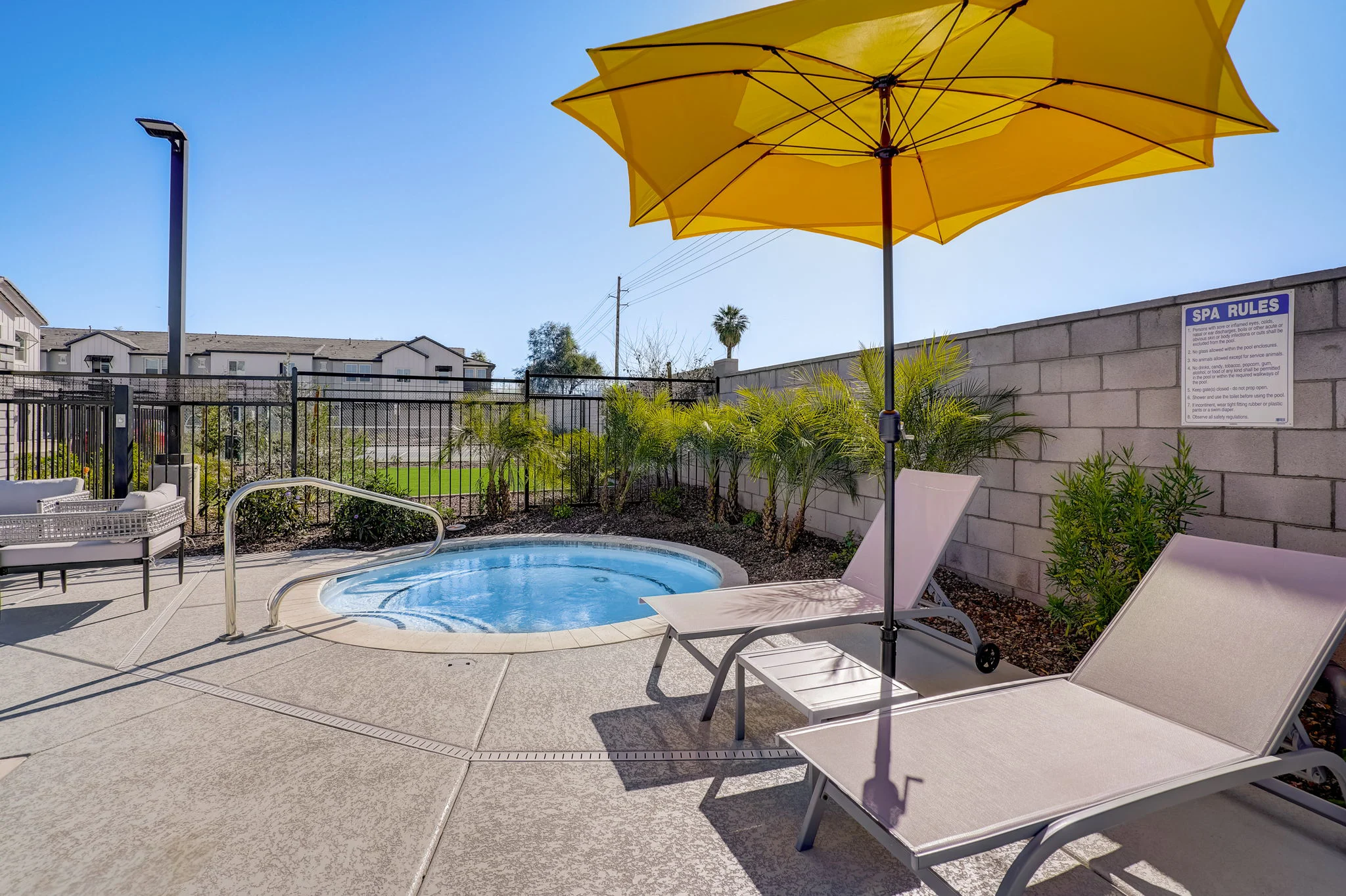 A spa area featuring a small hot tub with a yellow umbrella and lounge chairs nearby, surrounded by green plants and a fence.