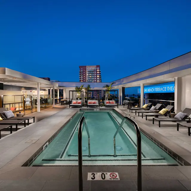 Rooftop Pool with City Views at Night A rooftop pool area at night with sleek modern design, featuring lounge chairs and palm trees, overlooking a city skyline.