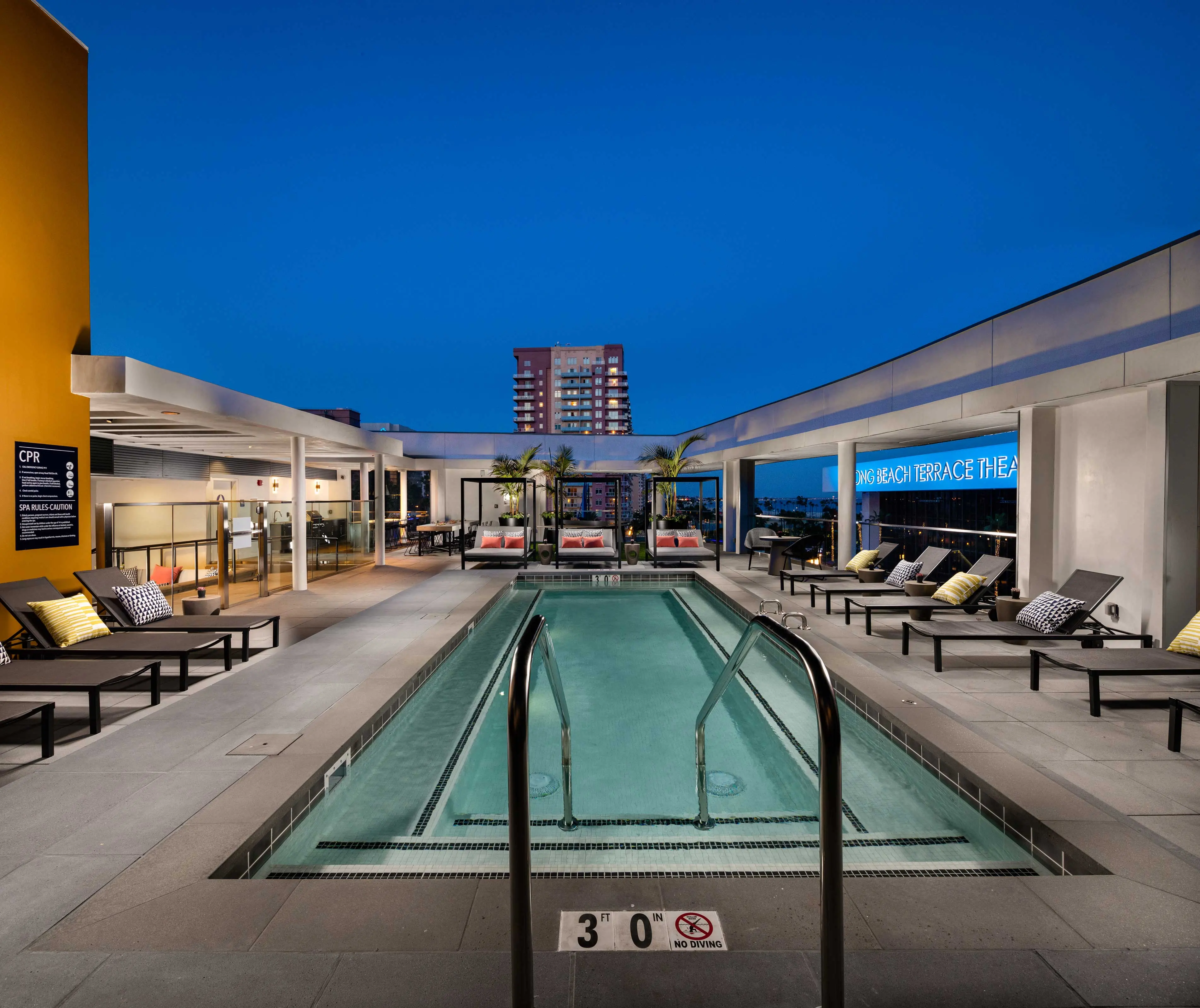A rooftop pool area at night with sleek modern design, featuring lounge chairs and palm trees, overlooking a city skyline.