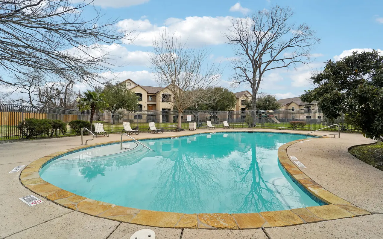 A clear swimming pool surrounded by a concrete deck, lounge chairs, and trees, with apartment buildings visible in the background under a blue sky.