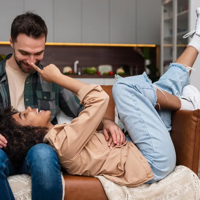 Couple in Love A couple relaxing on a sofa. The woman is lying with her feet up and playfully touching the man's face while he smiles at her. They are in a cozy living room setting.