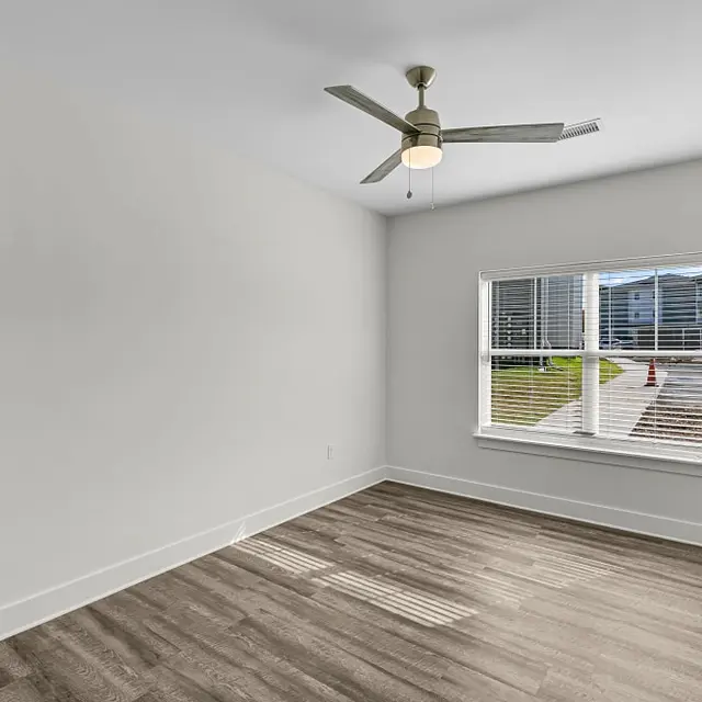 A spacious empty room with light wood flooring and a ceiling fan. There is a large window allowing natural light, and a door on the right side.
