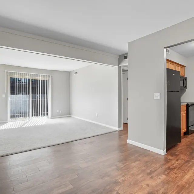 A spacious living area with a sliding glass door leading to a patio, featuring light-colored walls and wooden flooring. The adjoining kitchen appears to have dark appliances and wooden cabinets.