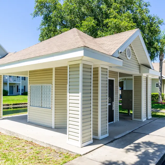 A small, enclosed mail kiosk with a peaked roof, situated in a green park-like area. The kiosk features a row of mailboxes on one side and is surrounded by grass and palm trees.
