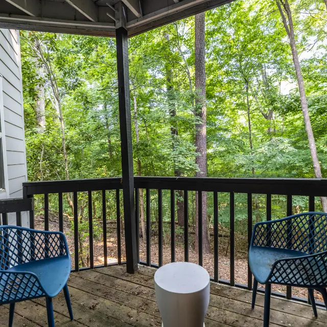 A cozy balcony with two blue chairs and a white table, surrounded by lush green trees.