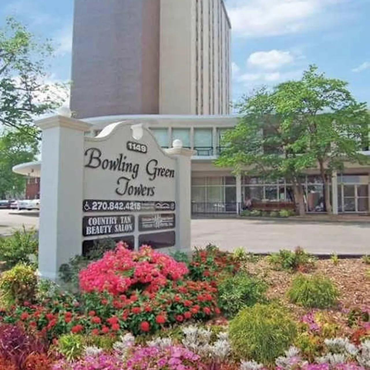 A sign for Bowling Green Towers surrounded by colorful flower beds in front of a tall building.