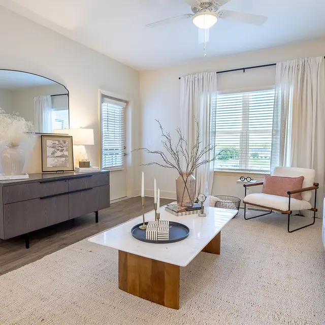 A modern living room featuring a light color palette with a white sofa, wooden coffee table, and decorative items. A large window allows natural light to enter, complemented by curtains. There's a stylish chair and a console table with a mirror reflecting the room's aesthetics.