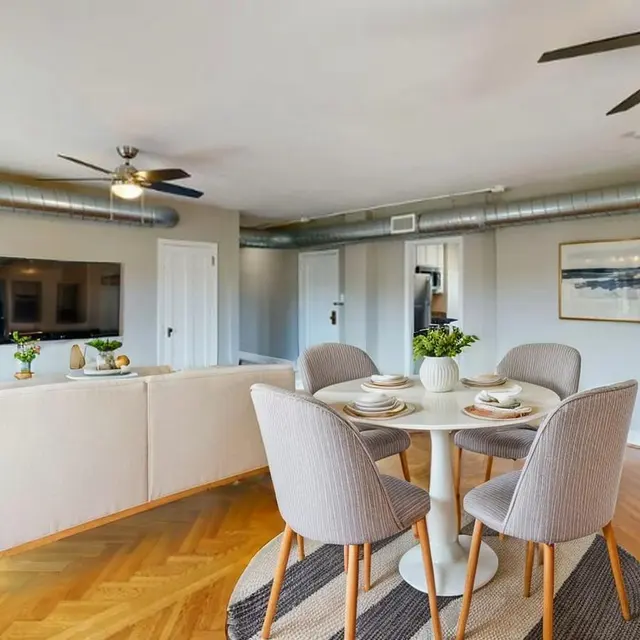 Bright and spacious modern apartment interior featuring a dining area with a round table and light-colored chairs, a comfortable sofa, and decorative elements. The space includes an entertainment unit and large windows for natural light.