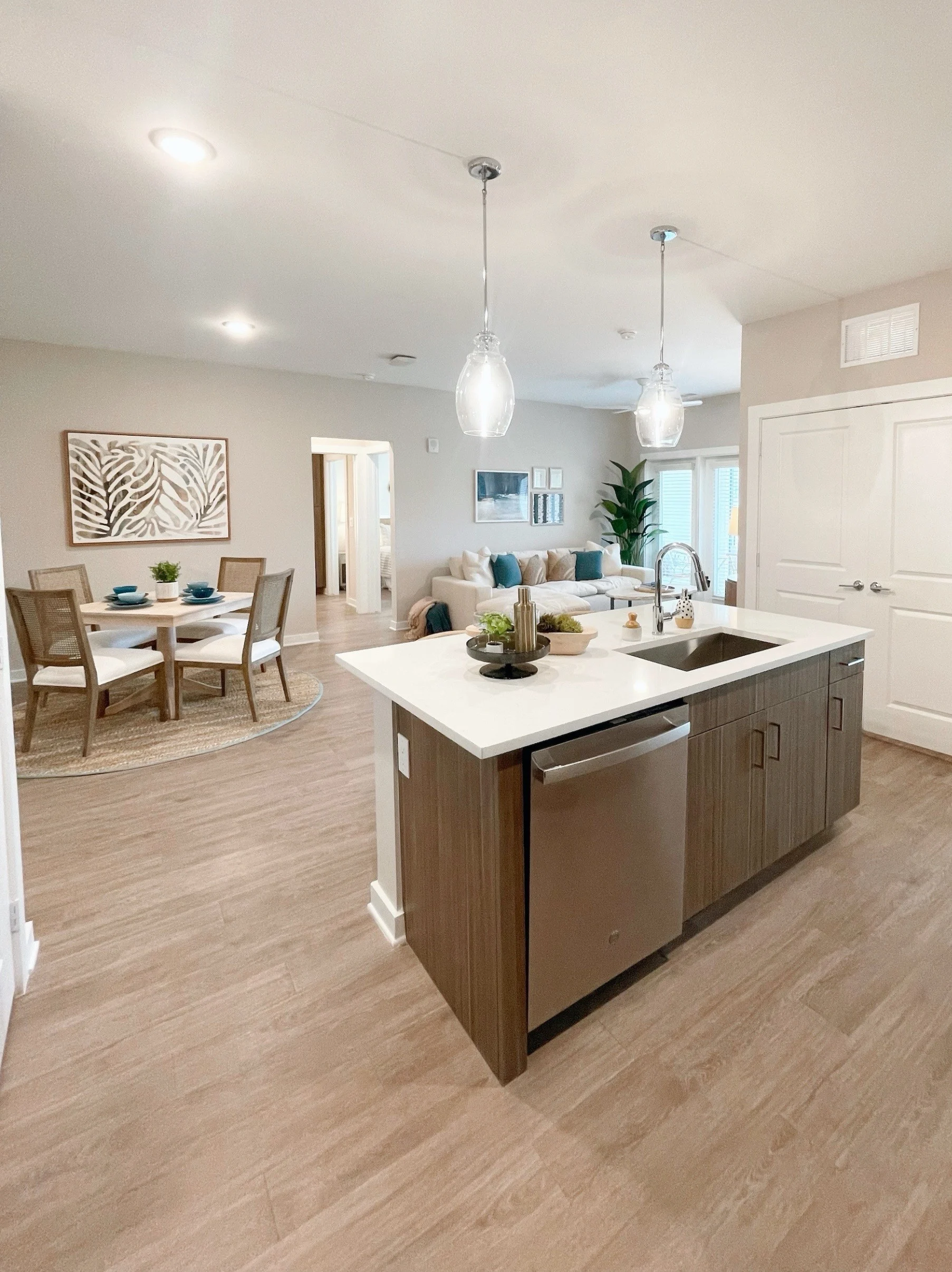 An open-concept kitchen with a modern design featuring a large island, wood cabinetry, and stainless steel appliances. The background shows a dining area and a cozy living room with soft furniture and large windows allowing natural light.