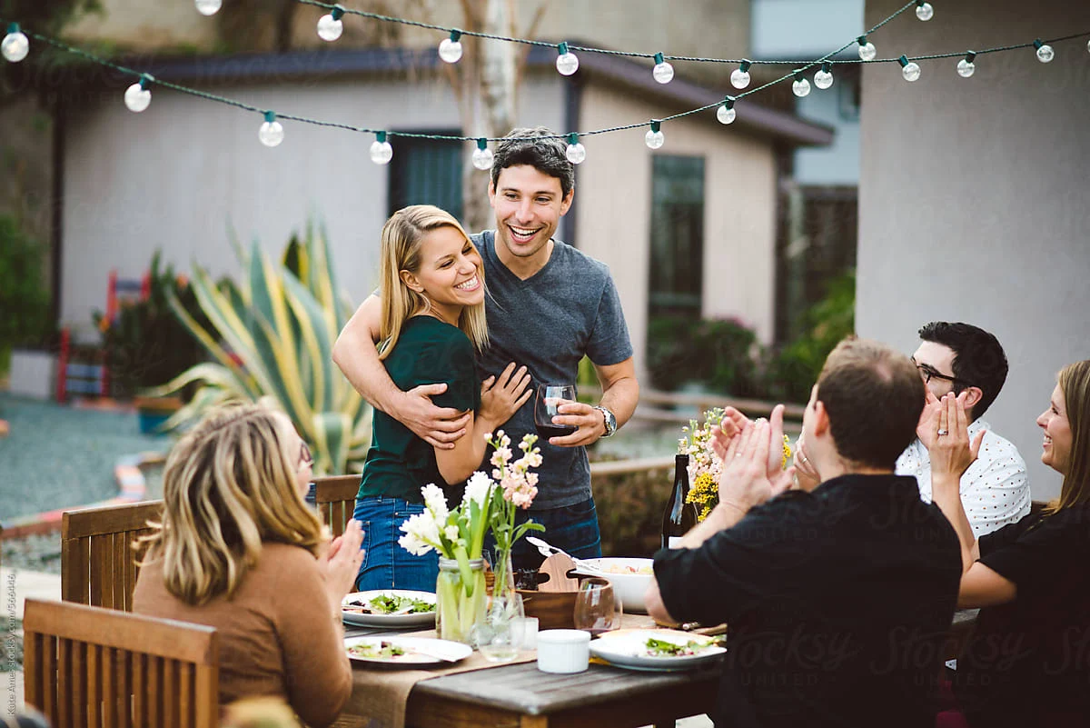 The Easton A lively outdoor dinner party scene with a group of friends celebrating. A couple is embracing, laughing, and holding drinks, surrounded by attendees at a table with plates of food. String lights decorate the setting, and there are plants in the background.