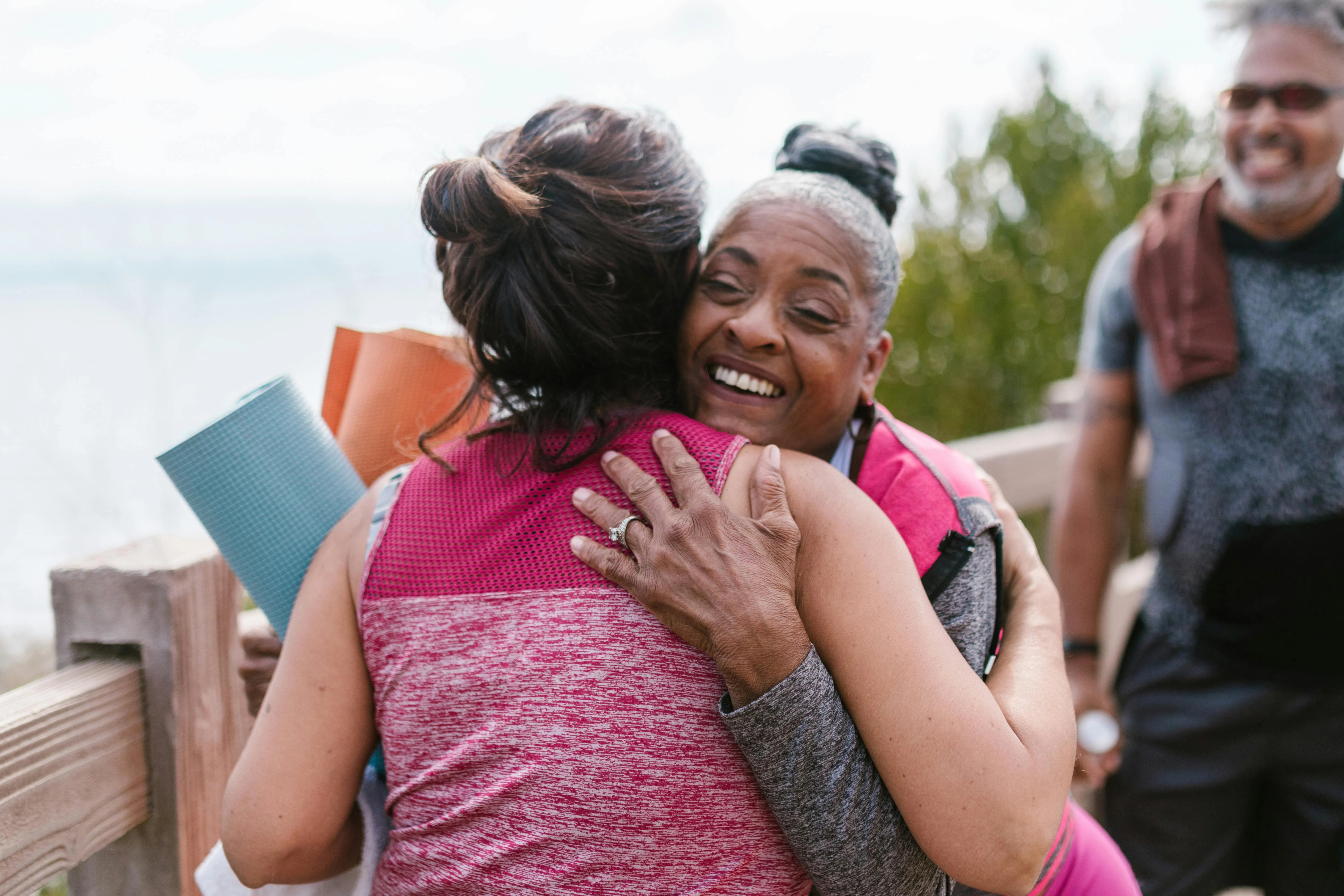 Warm Embrace Outdoors Two women embracing outdoors with smiles, one holding yoga mats. A man appears in the background.