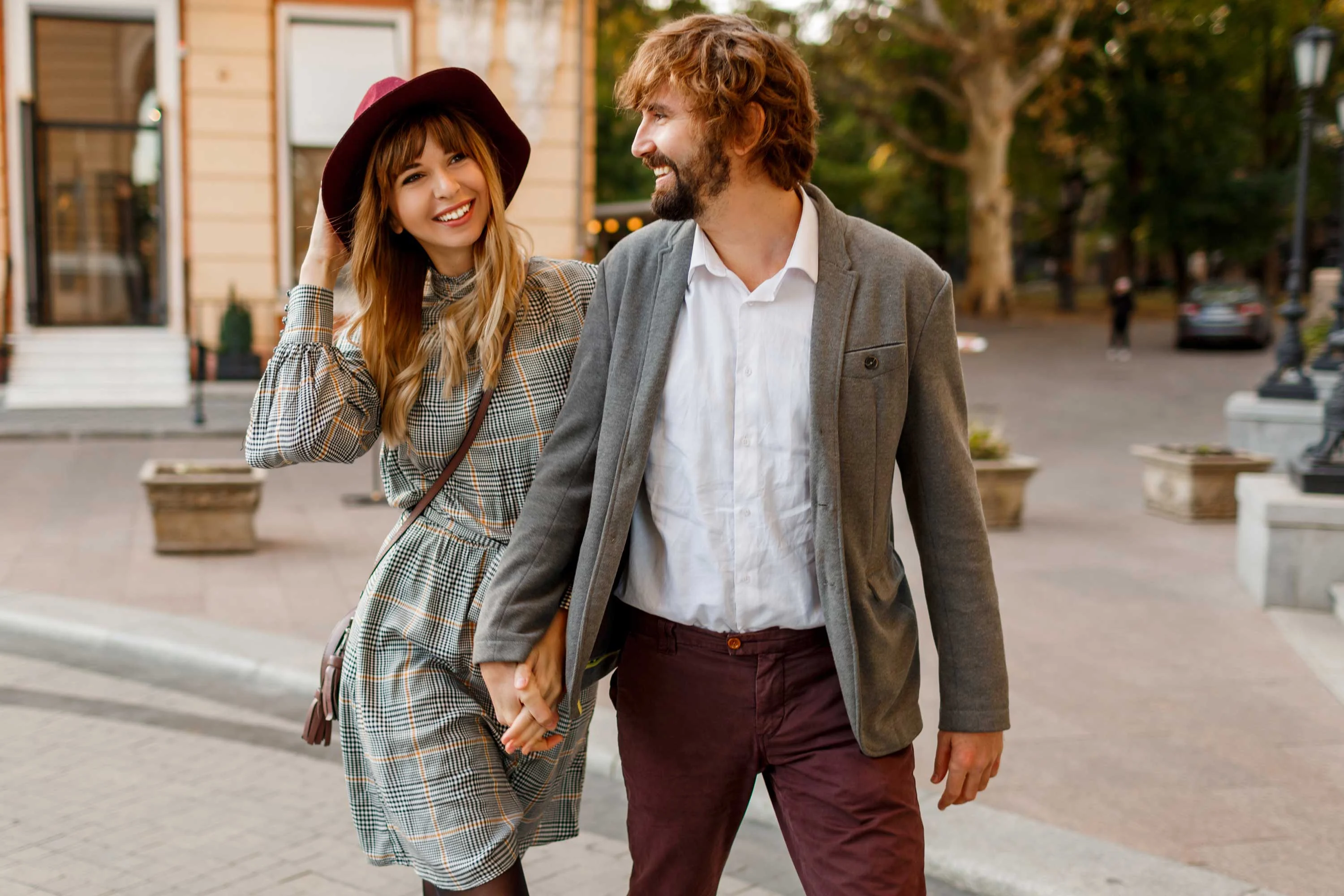 A couple walking hand in hand in an outdoor setting, dressed in stylish attire. The woman is wearing a patterned dress with a sweater and a burgundy hat, while the man is in a shirt and blazer with dark pants. They are smiling and looking at each other.