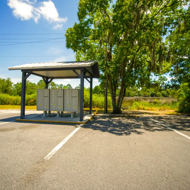 A postal mailbox stand with multiple compartments in a parking area, shaded by a tree.
