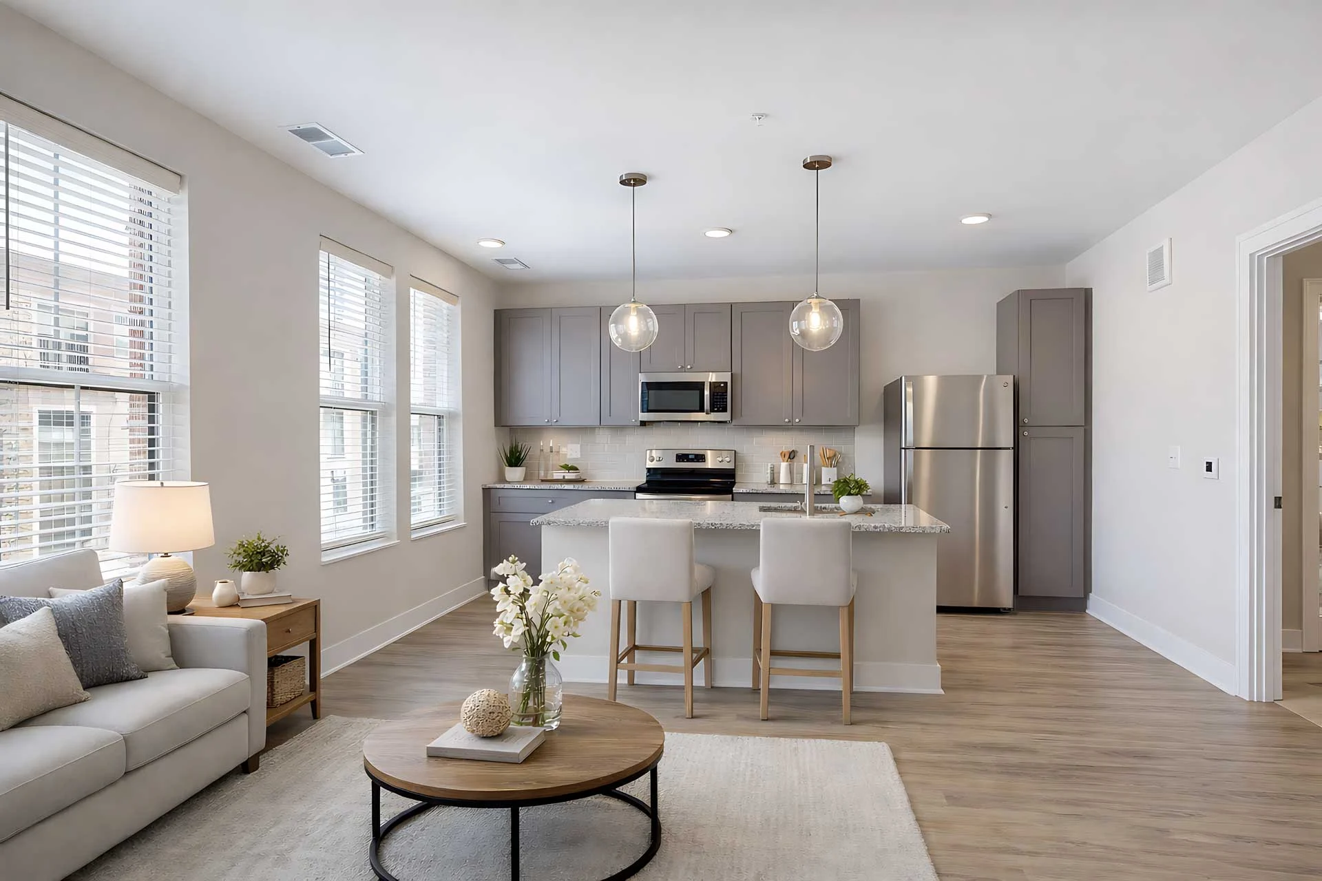 A modern kitchen and living room area featuring a light color scheme, with stainless steel appliances, pendant lighting, and large windows.