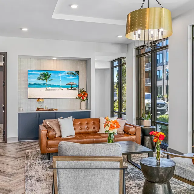 A modern lobby with large windows, a tan leather sofa, and a contemporary chandelier. Decorated with flowers and a beach-themed artwork.
