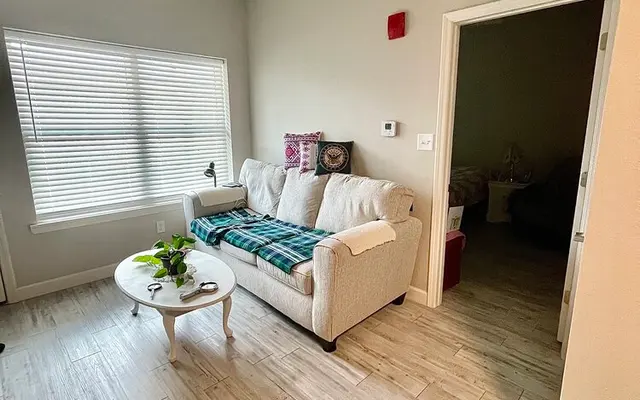 A cozy living room featuring a light-colored sofa with a blue blanket draped over it, a round coffee table, and a window with blinds. There is a ceiling fan above and a doorway leading to another room in the background.