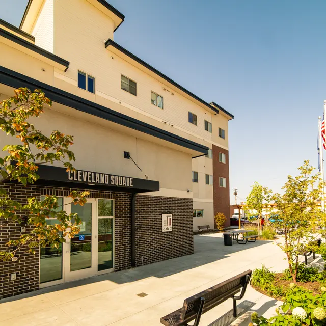 Cleveland Square Building Exterior view of Cleveland Square, a modern building with a sidewalk, bench, and greenery in the foreground, against a clear blue sky.