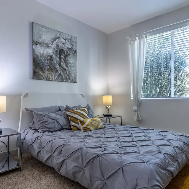 A cozy bedroom featuring a neatly made bed with gray linens and a decorative pillow, two bedside tables with lamps, and a large window with sheer curtains allowing natural light, along with a piece of abstract art on the wall.