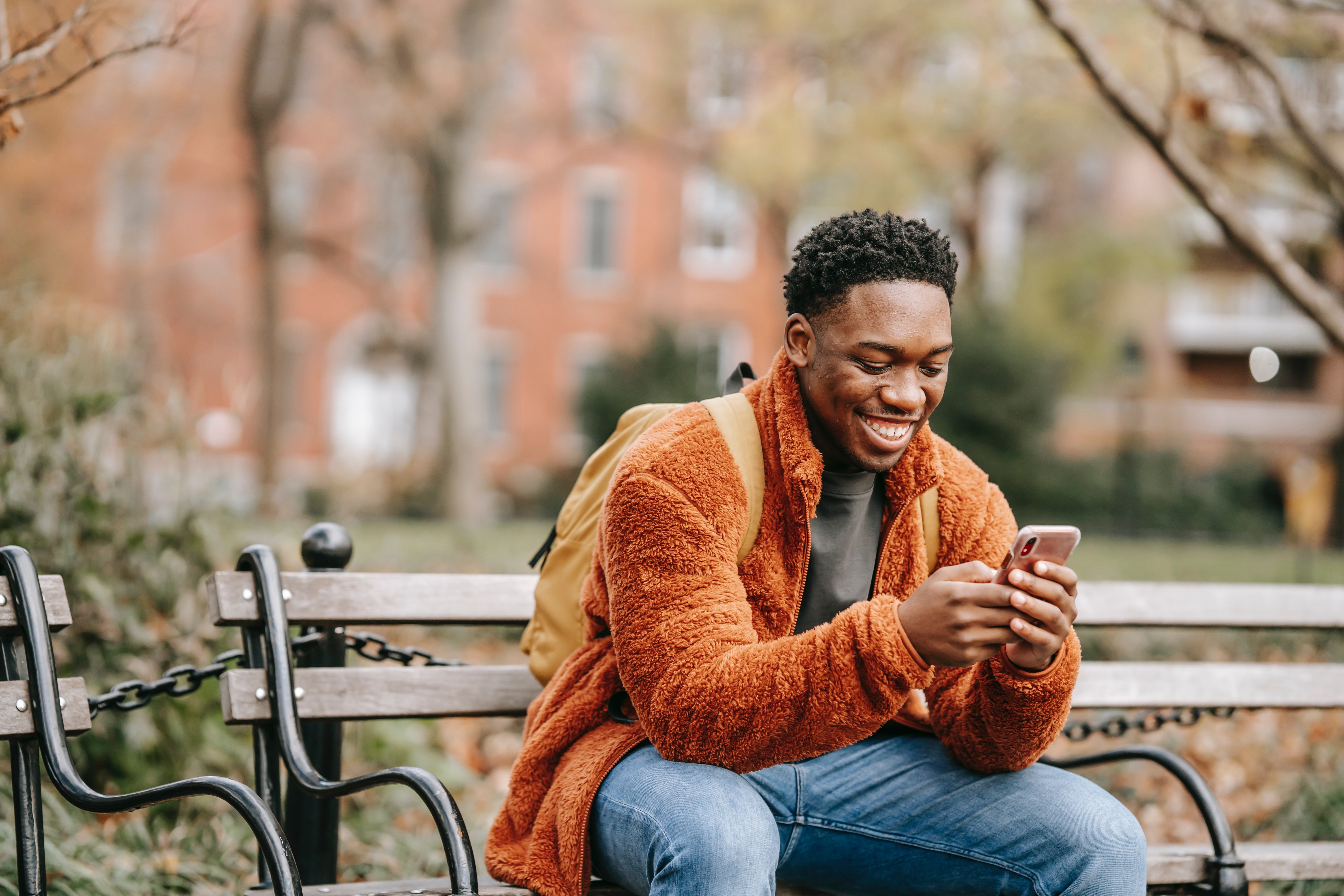 A young man sitting on a park bench, smiling while looking at his phone. He is wearing a cozy orange jacket and has a backpack beside him. The background features trees and buildings in an autumn setting.