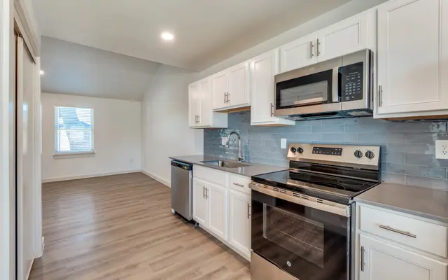 Modern Kitchen Design A modern kitchen with white cabinetry, stainless steel appliances, and a sleek countertop. The kitchen features a microwave and an oven, with a dishwasher visible next to the sink. Natural light enters through a window in the background.