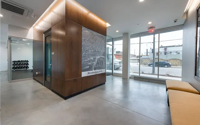 Modern Fitness Center Lobby A modern fitness center interior featuring a welcoming lobby with wooden paneling, large windows allowing natural light, a small seating area, and a view of weightlifting equipment behind glass.
