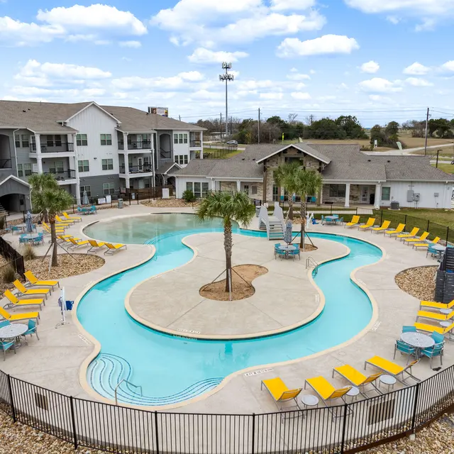 Aerial view of a resort-style swimming pool surrounded by lounge chairs and palm trees, with a nearby apartment complex in the background.