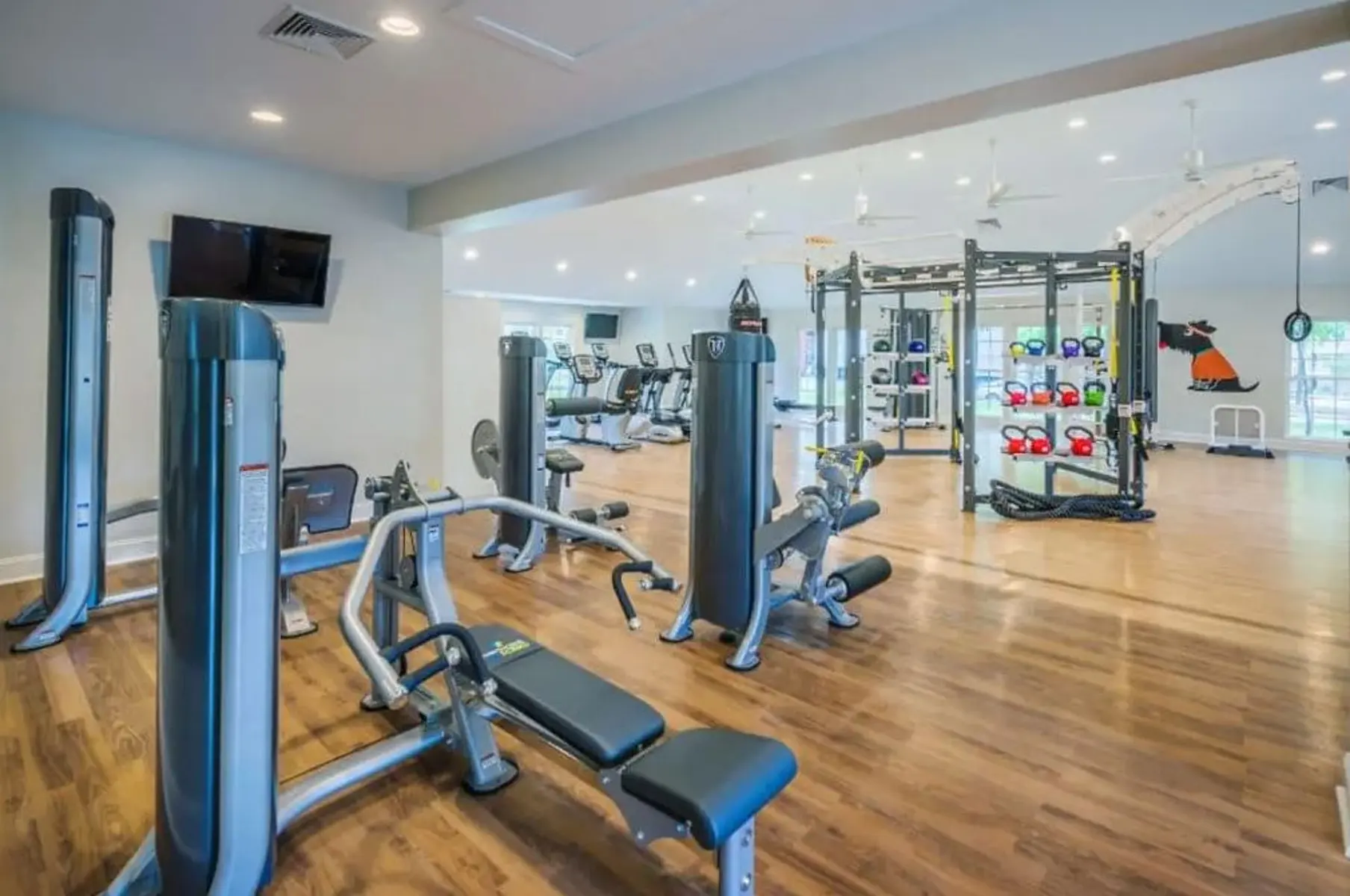 A spacious and modern gym interior featuring various exercise equipment, including weight machines, cardio machines, and a multi-functional weight training station. The room has wooden flooring and large windows allowing natural light in.