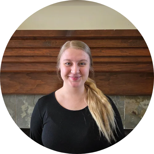 A young woman with long blonde hair, wearing a black top, smiling in front of a wooden mantel backdrop.