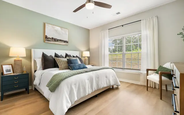 A well-decorated bedroom featuring a cozy bed with decorative pillows, a green blanket, and a minimalistic light fixture. There are bedside lamps and a window with sheer curtains allowing natural light.