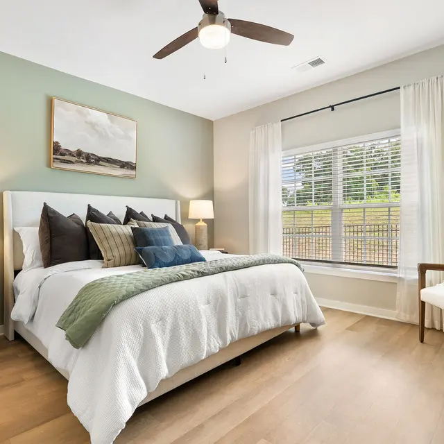 A well-decorated bedroom featuring a cozy bed with decorative pillows, a green blanket, and a minimalistic light fixture. There are bedside lamps and a window with sheer curtains allowing natural light.