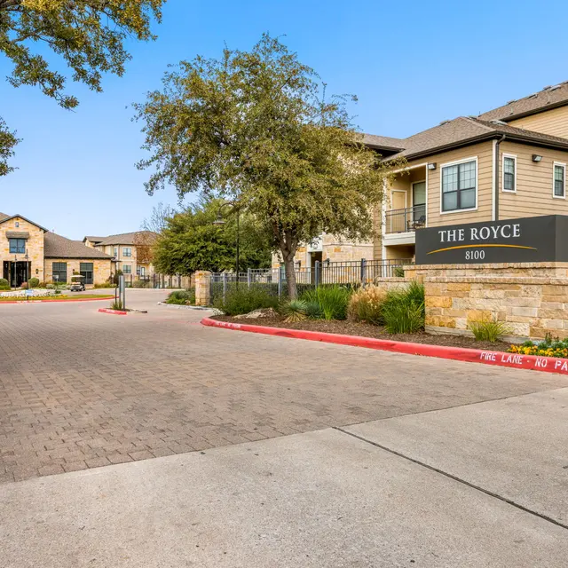 A view of the entrance to The Royce apartment complex, featuring a sign and landscaped surroundings with trees and flowers.
