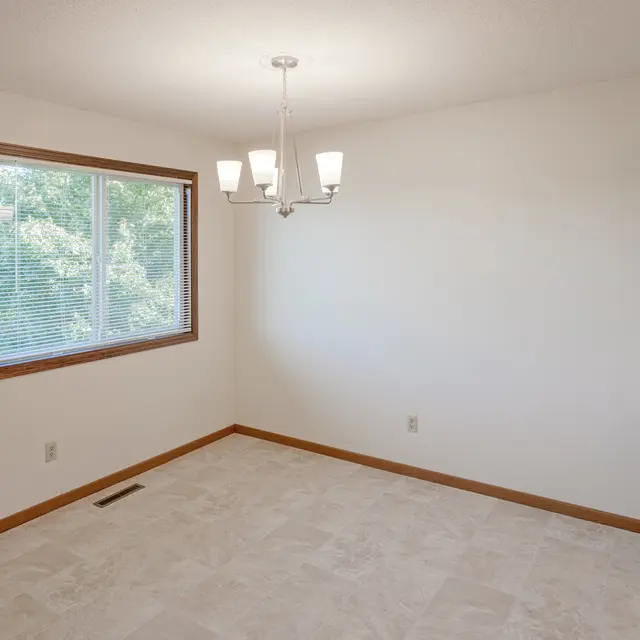 Court Place - Dining Room A bright, empty dining room featuring a large window with a view of greenery outside, beige tile flooring, and modern light fixture. There is a small mirrored cabinet on the right wall.