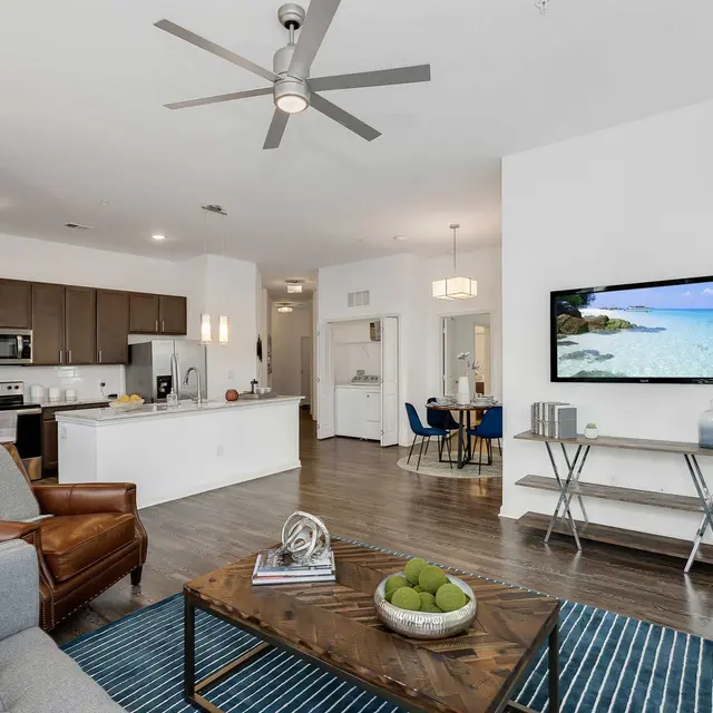 A contemporary living room featuring a spacious layout with a comfortable gray sofa, a brown leather chair, and a wooden coffee table with decorative items. The area has a sleek kitchen visible in the background with stainless steel appliances. A wall-mounted TV displays a beach scene, and there are accent tables and a modern ceiling fan present within the bright space.