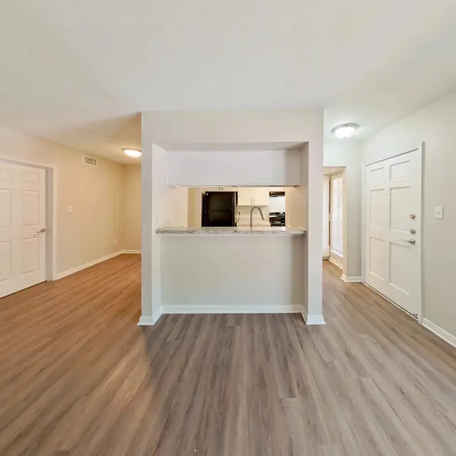 A spacious modern apartment interior featuring an open layout with wooden flooring, a small breakfast bar viewing into the kitchen, and large windows allowing natural light.