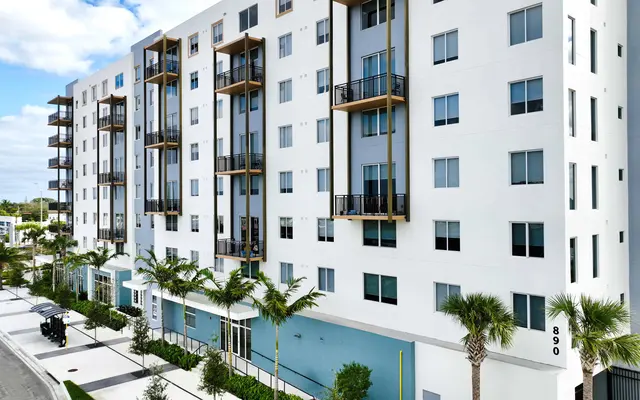 A modern apartment building with a white facade and large windows, featuring balconies and palm trees in front.