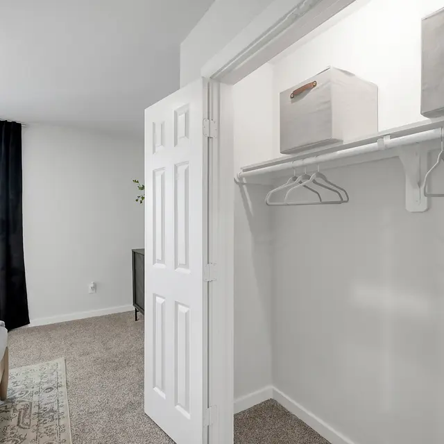 A modern bedroom featuring a neatly organized closet with storage boxes. There are two hangers and a bed with a patterned throw blanket in the foreground. A large window with black curtains allows natural light to enter the room.