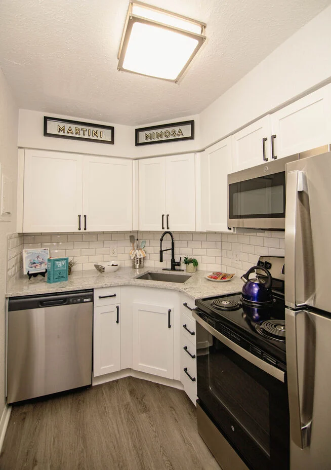 Stylish Corner Kitchen A modern corner kitchen featuring white cabinets, stainless steel appliances, and a beige tile backsplash. The decor includes wine-themed signs above the sink.