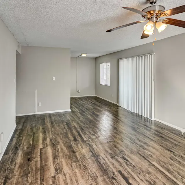 Interior view of a living room with wooden flooring, light-colored walls, and a ceiling fan. There are sliding glass doors on one side and a lit corner in the background.