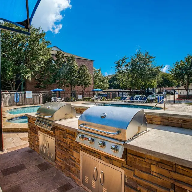An outdoor area featuring two stainless steel grills next to a pool, surrounded by trees and lounge chairs under a bright blue sky.