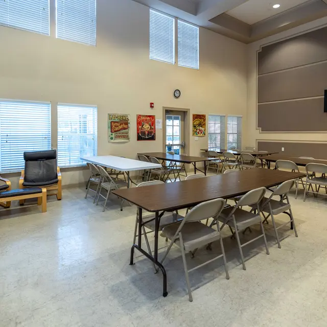 A spacious community room with several tables and chairs, two lounge chairs, and wall decorations featuring food and drink posters. A television is mounted on the wall in the background.