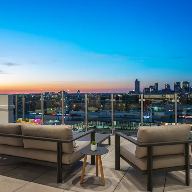 A rooftop lounge area featuring two modern sofas positioned on a patio with a glass railing, overlooking a city skyline at dusk.