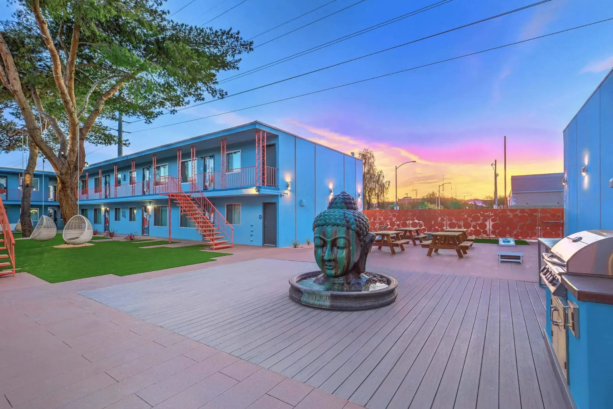 Outdoor community area featuring a decorative Buddha statue, picnic tables, and a barbecue grill, surrounded by bright blue apartment buildings under a colorful sky.