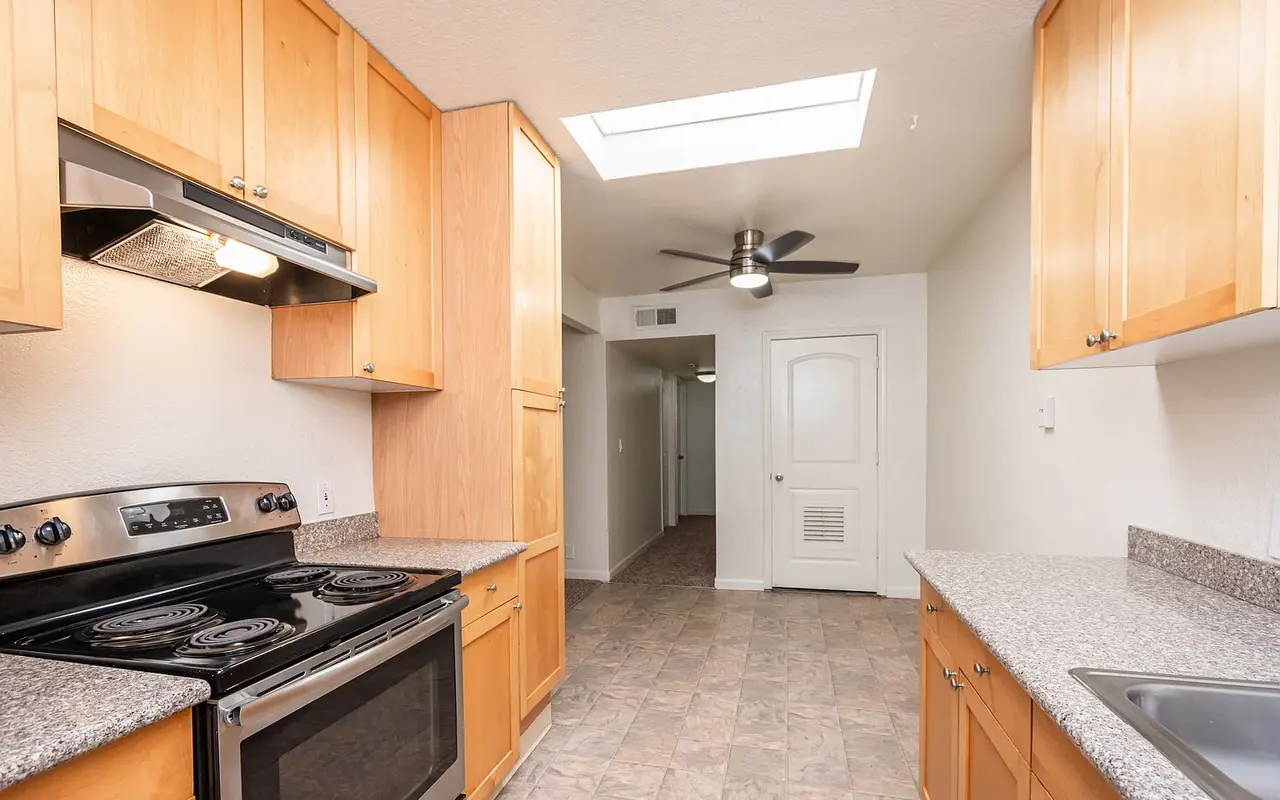 A modern kitchen featuring wooden cabinets, black stove, and granite countertops. The room is well lit with a skylight and includes a ceiling fan. A doorway can be seen in the background, leading to another room.