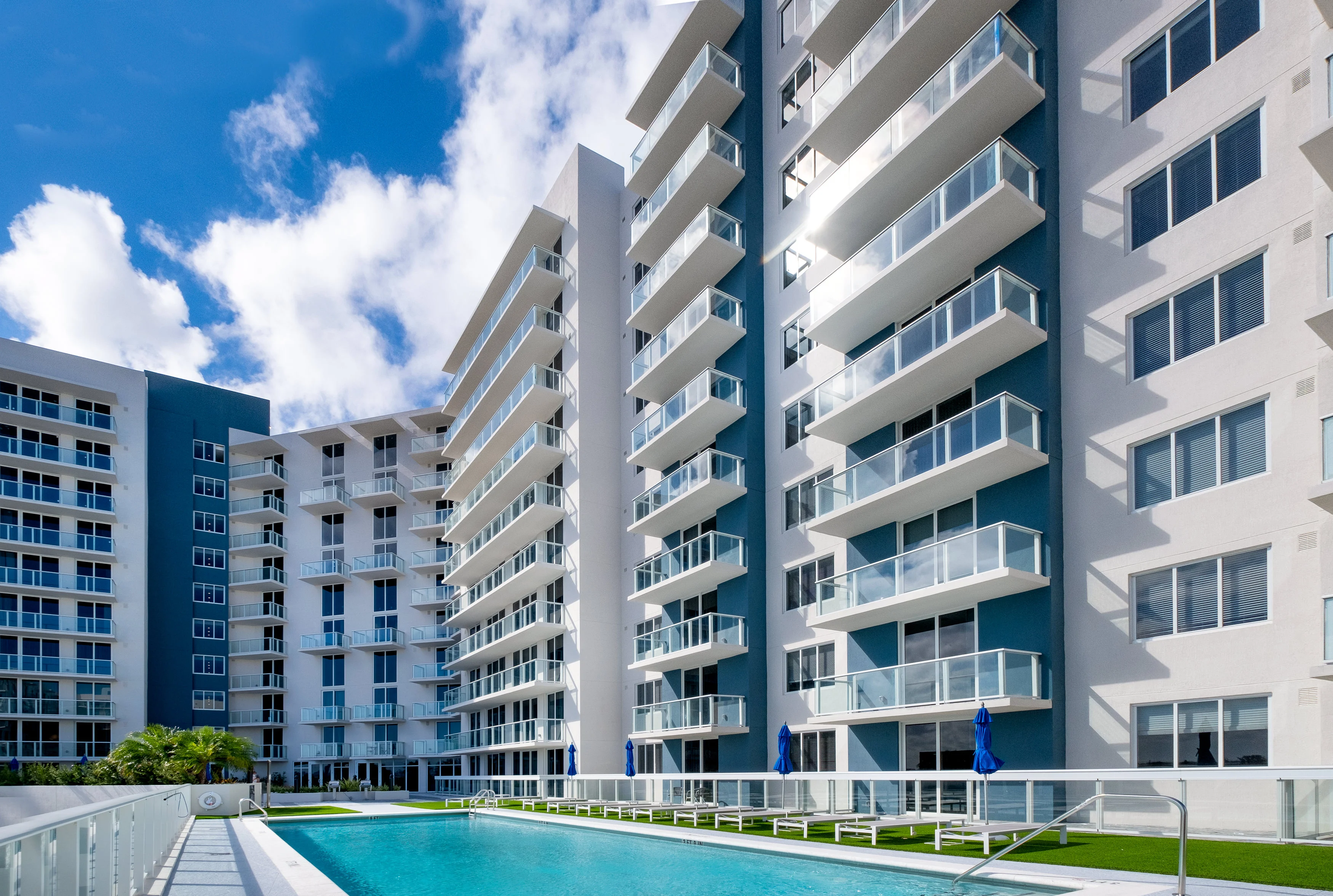 Modern Apartment Complex with Pool A modern apartment complex featuring multiple buildings with balconies and a swimming pool in the foreground under a blue sky.